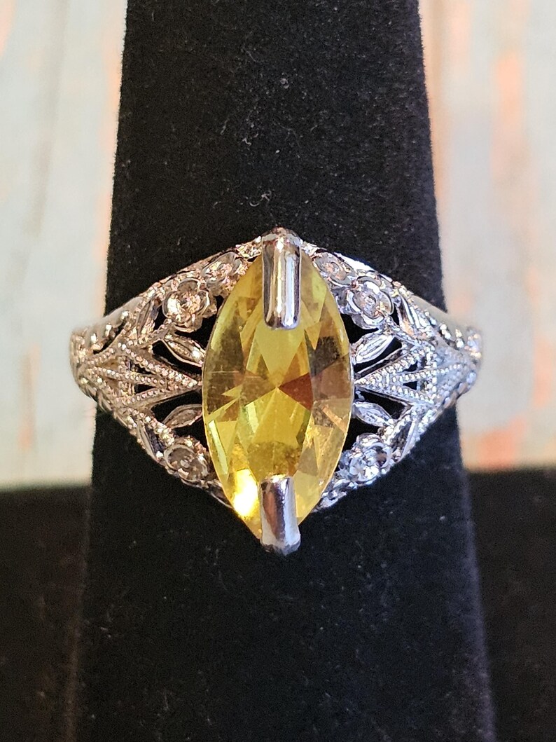 May include: A silver ring with a large, marquise-cut yellow gemstone. The ring features an ornate, filigree design with floral accents around the gemstone. The ring is displayed on a black stand.