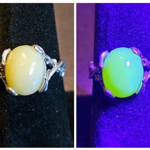 May include: A silver ring with a pale yellow oval gemstone set in a delicate, leafy design. The stone glows a bright green under a UV light.