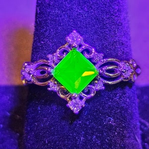 Könnte beinhalten: Ein silberner Ring mit einem grünen Edelstein, der in einer quadratischen Form gefasst ist. Der Ring hat ein dekoratives Filigranmuster und liegt auf einem violetten Samt-Hintergrund.