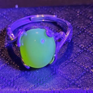 May include: A silver ring with a green gemstone. The ring is set in a delicate, floral design. The gemstone is glowing under a black light.