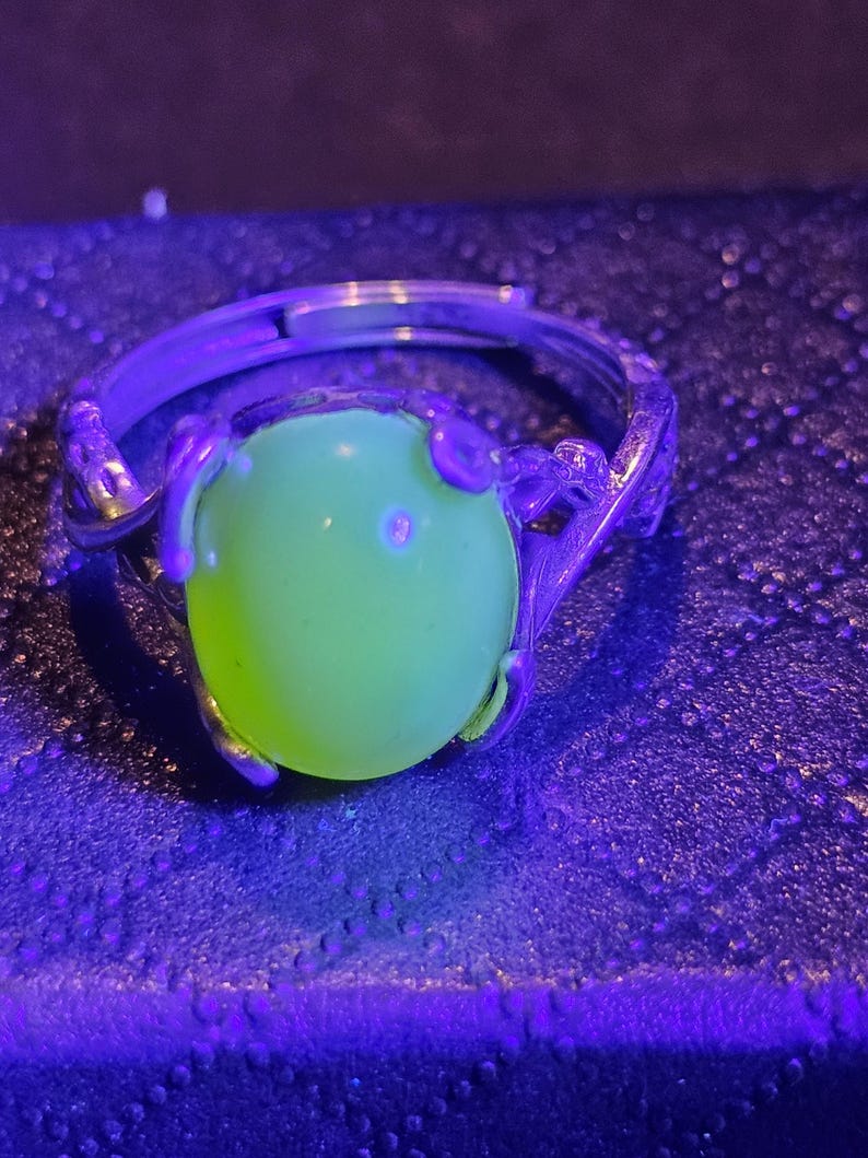 May include: A silver ring with a green gemstone. The ring is set in a delicate, floral design. The gemstone is glowing under a black light.