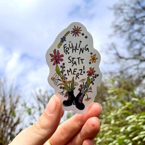 Könnte beinhalten: Ein Aufkleber mit dem deutschen Text "FRÜHLING STATT MERZ!" in Schwarz, umgeben von bunten Blumen und Grün. Das Design zeigt schwarze Stiefel, aus denen Blumen sprießen, vor einem bewölkten Himmel.