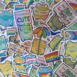 Könnte beinhalten: Eine Sammlung farbenfroher Froschaufkleber mit verschiedenen Designs und Texten. Einige Aufkleber zeigen Frösche, die Schilder mit Sätzen wie "My Gender is Cute", "Ribbit for Trans Rights" und "Hop for Equality!" halten. Andere haben Regenbögen und Herzen.