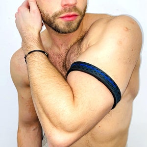 Puede incluir: Un hombre con el pelo corto castaño y barba lleva una pulsera de cuero negro con una raya azul. La pulsera está en su brazo izquierdo superior.