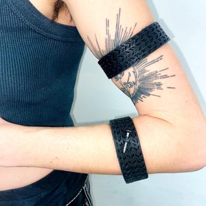 Può includere: Due braccialetti in pelle nera con un design testurizzato. I braccialetti sono indossati sul braccio superiore e sul polso.