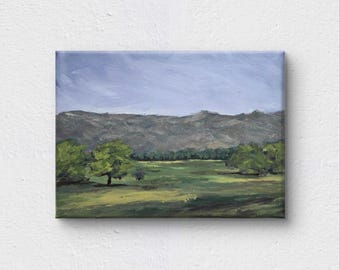 May include: An oil painting of a landscape scene. The artwork depicts a valley with green fields, trees, and a mountain range under a light blue sky. The painting is on a rectangular canvas.
