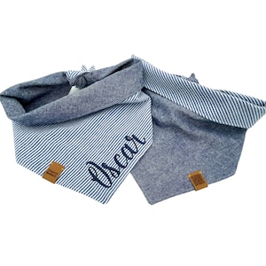 May include: Two blue dog bandanas. One is striped with the name "Oscar" embroidered in a cursive font. The other is a solid blue. Both bandanas have a brown leather tag with a logo. The bandanas are tied together.