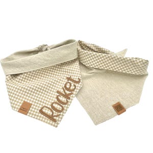 May include: Two beige dog bandanas. One features a tan and white gingham pattern, the name "Rocket" embroidered in brown, and a small brown leather tag. The other is solid beige with a brown leather tag.