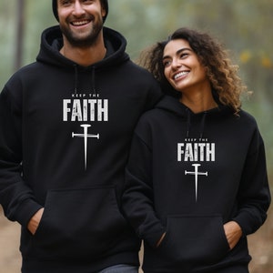 Op de afbeelding: Twee zwarte hoodies met de tekst "Keep the Faith" en een gestileerd kruis met een spijkerontwerp in wit.