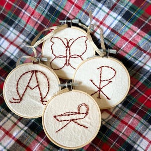 May include: Four small, round, wooden embroidery hoops with white fabric and red embroidery. The designs include a butterfly, a cross, a letter A, and a manger.
