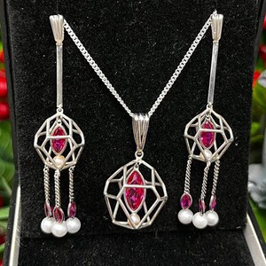 May include: A silver necklace and earrings set with a geometric cage design. Each piece features a pink gemstone and a pearl accent.