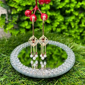 May include: Gold-toned drop earrings with geometric designs, red accents, and dangling pearls. The earrings are displayed on a round, mirrored surface with a crystal-studded border, set against a backdrop of green foliage.