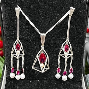 May include: A silver necklace and earrings set with a geometric design. The pendant and earrings feature a red gemstone surrounded by a silver cage with a pearl at the bottom. The earrings have three dangling pearls and red gemstones.