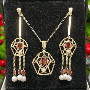 May include: A gold necklace and earrings set with a geometric pendant and dangling red gemstones. The pendant and earrings feature a cage-like design with a single red gemstone in the center.