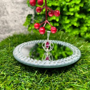 May include: A silver pendant with a geometric design featuring a red gemstone and a smaller white pearl. The pendant is reflected in a mirrored surface.