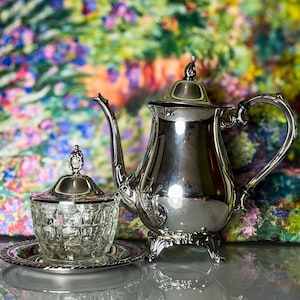 May include: A silver tea set with a teapot, sugar bowl, and a small tray. The teapot has a curved spout and handle, and sits on three small feet. The sugar bowl is clear with a textured design and a lid. The background is a colorful floral painting.
