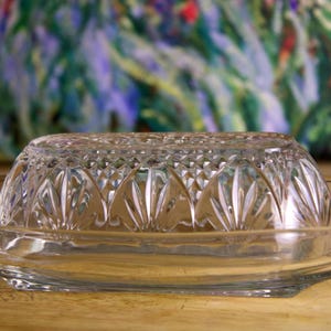 以下が含まれることがあります： ドーム型の蓋と浅い楕円形のベースを備えた、透明なプレスガラスのバター皿。 蓋には装飾的な繰り返し模様があります。 皿は木製の表面に置かれています。