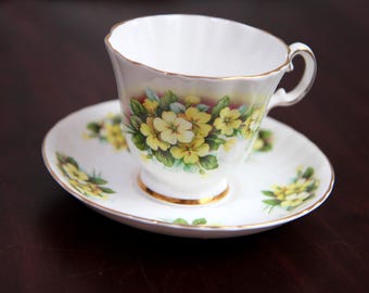 May include: White teacup and saucer set with a floral design featuring yellow flowers and green leaves. The rim of the cup and saucer are trimmed with gold. The teacup has a curved handle.
