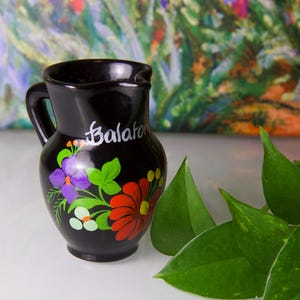 May include: Black ceramic pitcher with a handle and spout, featuring hand-painted floral designs in red, purple, and green. The word "Balaton" is written in white script on the side. A decorative piece for the home.
