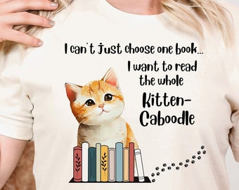 May include: A white t-shirt with a cute orange tabby cat sitting on a stack of colorful books. The text on the shirt reads "I can't just choose one book... I want to read the whole kitten-caboodle."