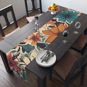 Op de afbeelding: Een bloemen tafelkleed met een beige achtergrond en kleurrijke bloemen in tinten van oranje, turquoise en rood. De tafelkleed ligt op een donkerhouten tafel met een set witte borden en bestek.