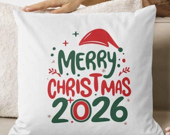 Może przedstawiać: Biała kwadratowa poduszka ozdobiona świąteczną grafiką z napisem Merry Christmas 2026 wykonanym czerwoną i ciemnozieloną czcionką. Wzór zawiera czerwoną czapkę Mikołaja umieszczoną nad tekstem, uzupełnioną małymi zielonymi i czerwonymi kropkami oraz gwiazdkami.