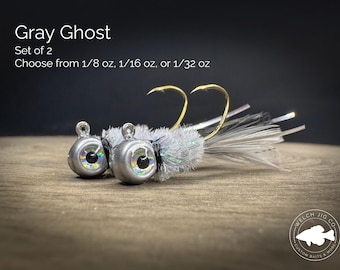 May include: Two Gray Ghost fishing jigs with silver heads, large holographic eyes, and white feathered tails. Each jig features a gold hook and is available in 1/8 oz, 1/16 oz, or 1/32 oz. The text reads Gray Ghost, Set of 2, Choose from 1/8 oz, 1/16 oz, or 1/32 oz.