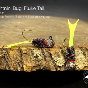 May include: Set of two black and red fishing lures with yellow fluke tails. The lures are on a wooden surface. The text "Lightnin' Bug: Fluke Tail" and "Set of 2" is above the lures. The text "Choose from 1/8 oz, 1/16 oz, or 1/32 oz" is below the lures.