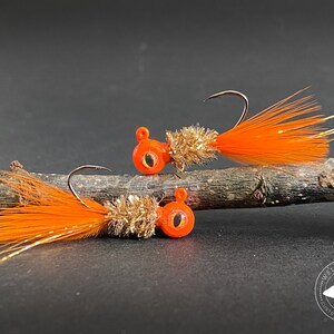 May include: Two orange and gold jig heads with orange feathers. The jig heads have a black eye and are on a brown branch. The text "Welch Jig Co. Custom Baits & More" is on the bottom right of the image.
