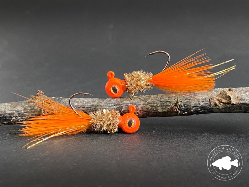 May include: Two orange and gold jig heads with orange feathers. The jig heads have a black eye and are on a brown branch. The text "Welch Jig Co. Custom Baits & More" is on the bottom right of the image.