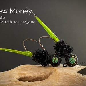 May include: Two black and green fishing jigs with green and yellow feathers. The jigs have a black and green eye design and are labeled "Welch Jig Co. Custom Baits & More".