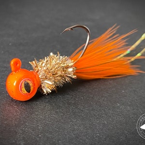 May include: Orange and gold fishing lure with a feathered tail and a round head with a black eye. The lure is on a gray surface with a logo that says "Welch Jig Co. Custom Baits & More".