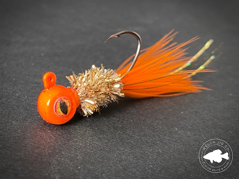 May include: Orange and gold fishing lure with a feathered tail and a round head with a black eye. The lure is on a gray surface with a logo that says "Welch Jig Co. Custom Baits & More".