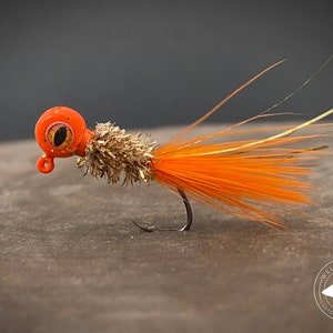 May include: Orange and brown fishing lure with a large eye and a long tail of orange feathers. The lure is on a brown surface with a logo that reads "Welch Jig Co. Custom Baits & More".