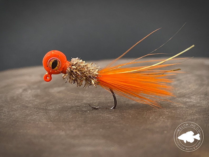 May include: Orange and brown fishing lure with a large eye and a long tail of orange feathers. The lure is on a brown surface with a logo that reads "Welch Jig Co. Custom Baits & More".