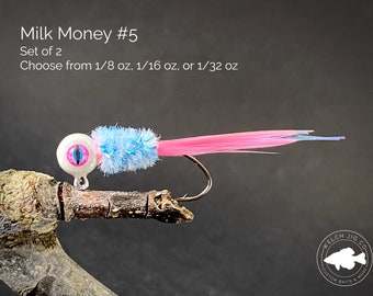 May include: A set of two pink and blue fishing lures with a white eye and a pink feather tail. The lures are called "Milk Money #5" and are available in 1/8 oz, 1/16 oz, or 1/32 oz.