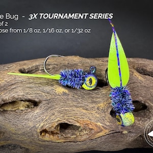 May include: Two June Bug 3X Tournament Series fishing jigs. One jig has a chartreuse blade with a blue stripe, and the other has a blue and yellow head with a chartreuse tail. The text reads "June Bug - 3X TOURNAMENT SERIES".