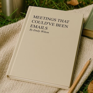 May include: A book titled "MEETINGS THAT COULD'VE BEEN EMAILS" by Emily Wilson, with a light beige cover, rests on a beige woven blanket. A stainless steel tumbler and a pencil are also present.
