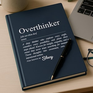 Navy blue hardcover journal titled “Overthinker” with a dictionary-style definition and personalized name on the cover, styled on a minimalist desk beside a coffee mug, laptop, pen, and glasses. A modern personalized overthinker notebook gift, perfect for journaling thoughts, stress relief, mental health support, and funny relatable gifts for coworkers or friends who overthink.