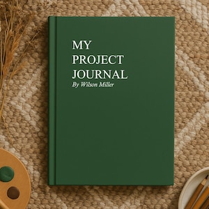 May include: A dark green journal with the text "MY PROJECT JOURNAL By Wilson Miller" is displayed on a textured, neutral-toned surface. A wooden palette with paint, a paintbrush, and colored pencils are arranged around the journal.
