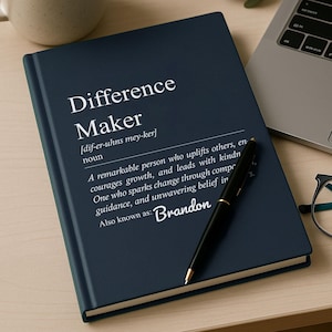 May include: A navy blue journal with the words "Difference Maker" printed on the cover, along with a definition. A black pen rests on the journal. A cup of coffee, a laptop, and glasses are also visible.