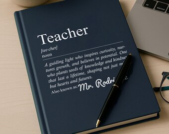 May include: A navy blue notebook with the word "Teacher" in white, along with a definition. A black pen rests on the notebook. A cup of coffee, a laptop, and glasses are also on the wooden surface.