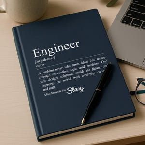 May include: A navy blue notebook with the word "Engineer" in white, along with a definition. A black pen rests on the notebook, next to a laptop, a cup of coffee, and glasses.