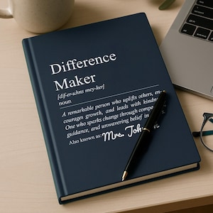 May include: A dark blue hardcover notebook featuring the white printed text Difference Maker, a phonetic pronunciation, a definition, and the name Mrs. Johnson. A black ballpoint pen rests diagonally across the front cover of the notebook.