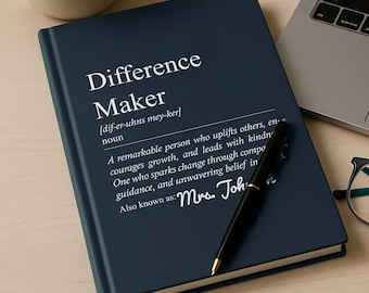 May include: A dark blue hardcover notebook featuring the white printed text Difference Maker, a phonetic pronunciation, a definition, and the name Mrs. Johnson. A black ballpoint pen rests diagonally across the front cover of the notebook.