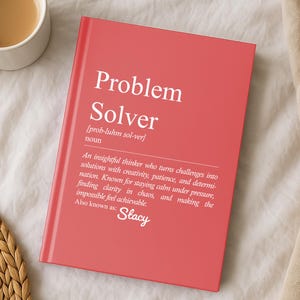 May include: A coral-colored notebook with the words "Problem Solver" in white. The definition of "Problem Solver" is printed below the title. A cup of coffee sits to the left of the notebook.