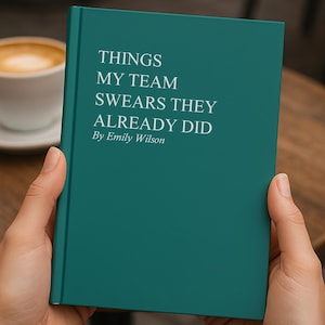 May include: A teal book titled "THINGS MY TEAM SWEARS THEY ALREADY DID" by Emily Wilson. The book is held in front of a blurred background with a cup of coffee on a table.
