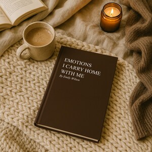 May include: A brown book titled "EMOTIONS I CARRY HOME WITH ME" by Emily Wilson rests on a cream-colored knitted blanket. A cup of coffee and a lit candle are also present, creating a cozy, warm atmosphere. An open book is visible in the background.