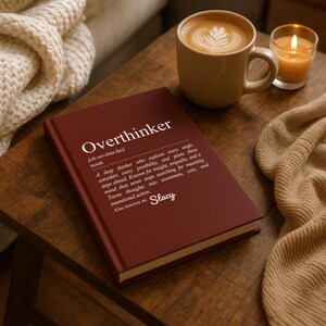 May include: A maroon hardcover book titled "Overthinker" with a definition printed on the cover. A cup of coffee with latte art and a lit candle sit nearby on a wooden table. A cozy blanket is also present.