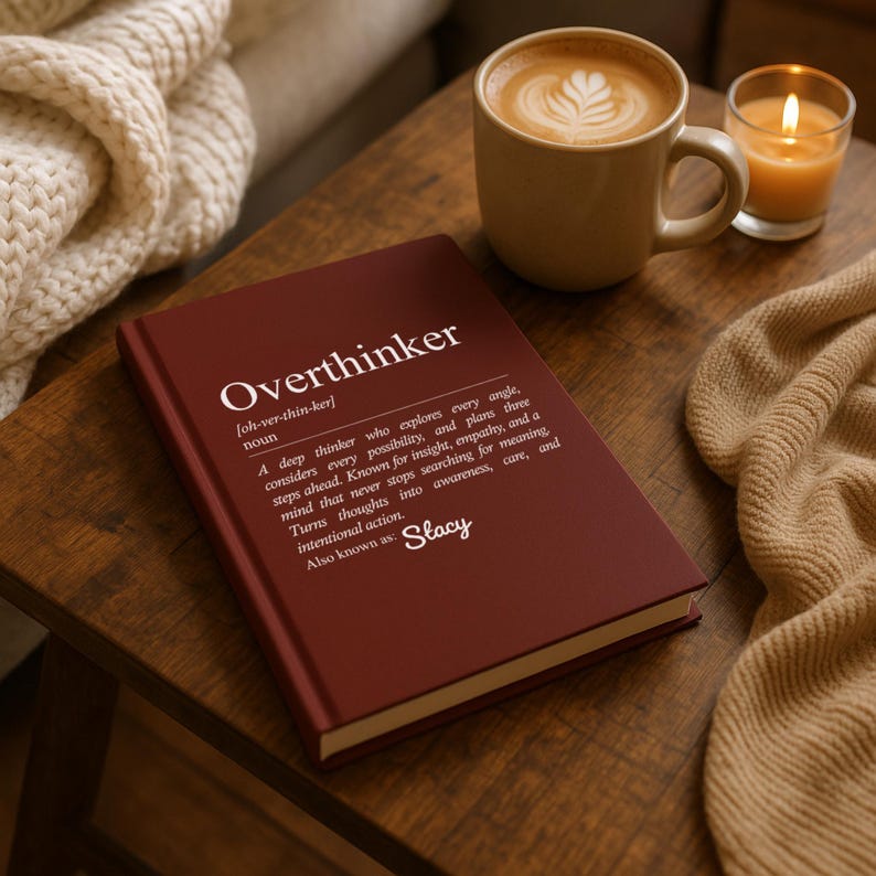May include: A maroon hardcover book titled "Overthinker" with a definition printed on the cover. A cup of coffee with latte art and a lit candle sit nearby on a wooden table. A cozy blanket is also present.